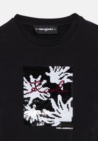 Μαύρο βαμβακερό μπλουζάκι με σχέδιο από τετράγωνα παγιέτες σε λευκό και μαύρο με κόκκινο κείμενο. Η ετικέτα αναγράφει "KARL LAGERFELD KIDS." Στρογγυλή λαιμόκοψη.