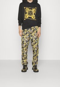 Černá mikina s kapucí s žlutým grafickým potiskem a šňůrkami; doplněná vzorovanými žluto-černými kalhotami. Nošena s černo-zlatými botami.