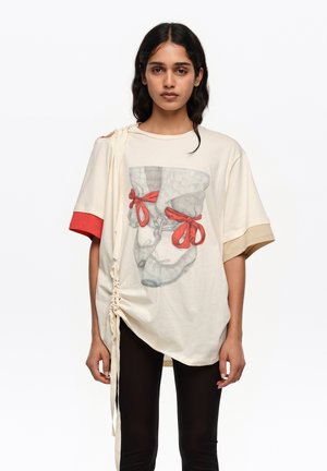 Kobieta w oversize'owej kremowej koszulce z czerwono-beżowymi rękawami, z nadrukiem baletowego bucika z czerwonymi kokardkami i marszczonym trokiem z boku, w połączeniu z czarnymi legginsami.