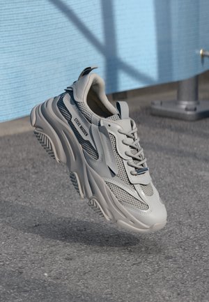 Hellgrauer klobiger Sneaker mit Mesh-Einsätzen und Schnürsenkeln, Marke "Steve Madden", über Asphaltfläche nahe einer hellblauen Wand aufgehängt.