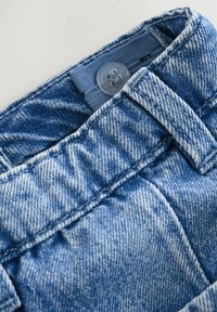 Primer plano de la cinturilla de unos pantalones vaqueros azules con un botón y un lazo para cinturón, con textura de tela y costura visibles.