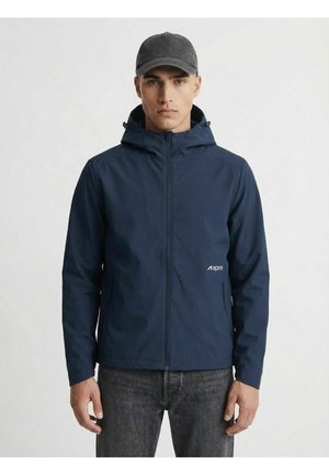 Hombre con chaqueta con capucha azul marino y gorra negra, de pie con las manos relajadas a los lados sobre un fondo blanco liso.