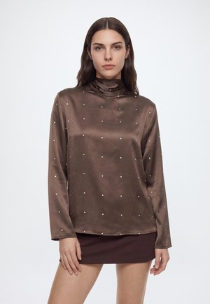 Femme portant un chemisier en satin marron à manches longues avec de petits pois blancs et une mini-jupe marron foncé sur un fond uni.