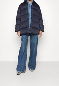 Daunenjacke in Dunkelmarineblau mit horizontalen Steppnähten, großem Kragen und Knopfverschluss, getragen über einem Denim-Hemd und ausgestellten Jeans.