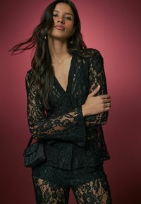 Black lace top with bell sleeves, v-neckline, textured pattern, paired with matching lace trousers and a small black handbag.
