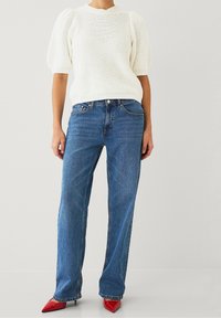 Maglione bianco a maglia con maniche a sbuffo, abbinato a jeans a gamba dritta blu e tacchi a punta rossi. Design e texture minimaliste.