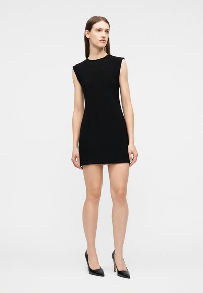 Γυναίκα στέκεται φορώντας μαύρο μίνι φόρεμα χωρίς μανίκια και μαύρα ψηλοτάκουνα παπούτσια μπροστά σε απλό φωτεινό φόντο.