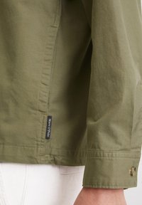 Olijfgroene jas met een gladde textuur, knoopdetail op de mouw en een zijkleurige label met "Marc O'Polo".