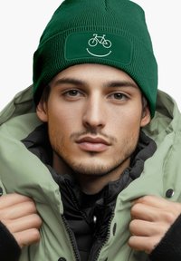 Green knit beanie with a smiley face and bicycle graphic, worn with a puffy, light green jacket. Smooth texture and snug fit.