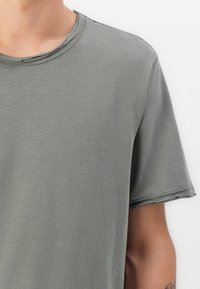 Kurzarm-T-Shirt mit Rundhalsausschnitt in gedämpftem Grün, mit weichem Stoff und rohem Kantenfinish am Ausschnitt und an den Ärmeln.