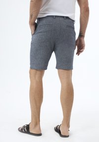 Shorts bleu chiné avec une coupe ajustée, devant plat et deux poches arrière. Portés avec des sandales noires, mettant en valeur une texture de tissu lisse.