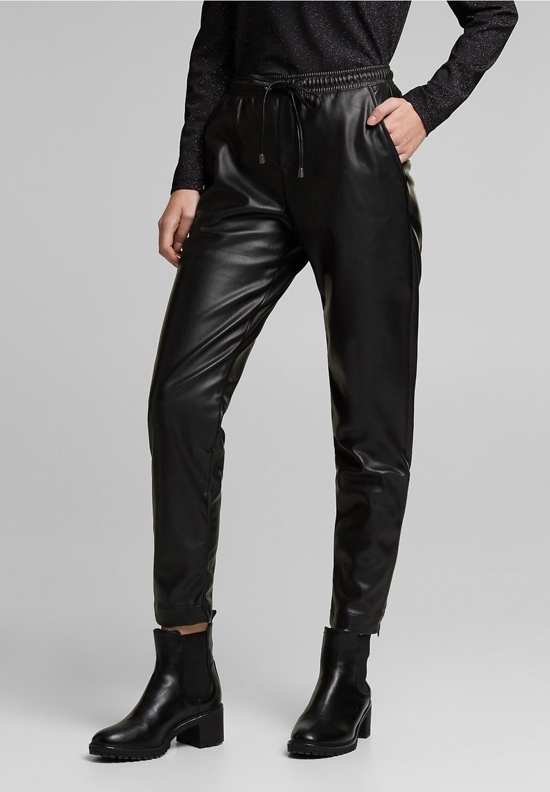 Nainen, joka käyttää mustia tekonahkahousuja, joissa on joustava vyötärönauha ja kiristysnauha, yhdistettynä mustiin nilkkureihin, seisoo harmaalla taustalla.