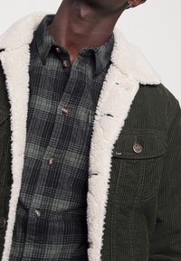 Green corduroy jacket with white faux fur lining, featuring a checked flannel shirt underneath and metal branding detail on the pocket.