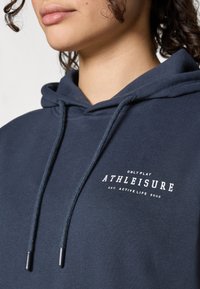 Námornícky modrá mikina s kapucňou s viazacou šnúrkou, jemnou textúrou a bielym logom "ATHLEISURE" na prednej strane.