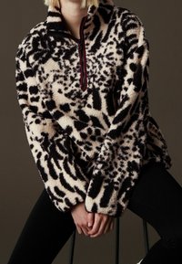 Osoba siedząca, ubrana w polar z czarno-białym wzorem, z kołnierzem na suwak do ćwiartki oraz czarne spodnie, na neutralnym tle.