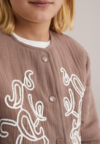 Prim-plan al unei persoane care poartă o jachetă deschisă la culoare cu nasturi, având broderie texturată albă și o cămașă albă cu guler în dungi dedesubt.