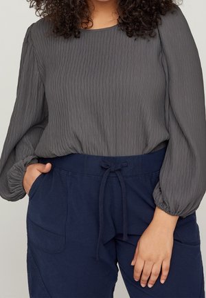 Mujer con blusa gris texturizada de manga larga metida en pantalones azul marino con cordón, con las manos en los bolsillos, cabello rizado visible.