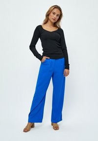 Zwart lange mouwen top met een ronde halslijn, gecombineerd met felblauwe wijde broekspijpen. Model draagt een paar beige blokhak sandalen. Effen achtergrond.