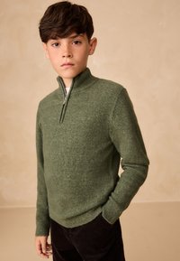 Grøn strikket sweater med halv lynlås i kraven og ribstrikkede manchetter, kombineret med mørke bukser i manchesterstof. Tekstureret, blødt udseende.