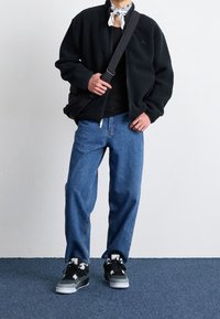  Czarna polarowa kurtka z wysokim kołnierzem, zestawiona z niebieskimi, luźnymi dżinsami oraz czarnymi tenisówkami z szarymi akcentami i białym logo.