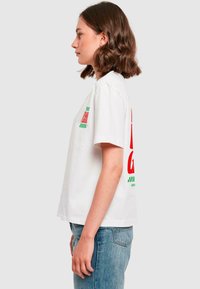 T-shirt en coton blanc avec des manches courtes et une coupe carrée. Le devant, au niveau de la poche gauche, présente un texte graphique rouge et vert, agrémenté d'un motif en damier.