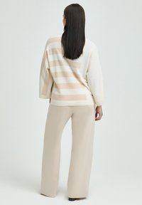 Übergroßer Pullover in Creme mit hellbeigen Streifen, weiche Textur, weite Ärmel, kombiniert mit fließenden beigen Hosen.