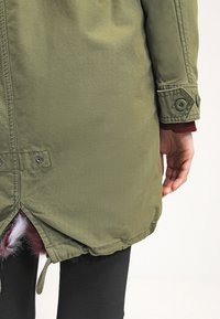 Oliwkowa parka o miękkiej fakturze, z detalami w postaci guzików i ściągaczem u dołu. Wyłożona sztucznym futrem przy otwarciu z tyłu.