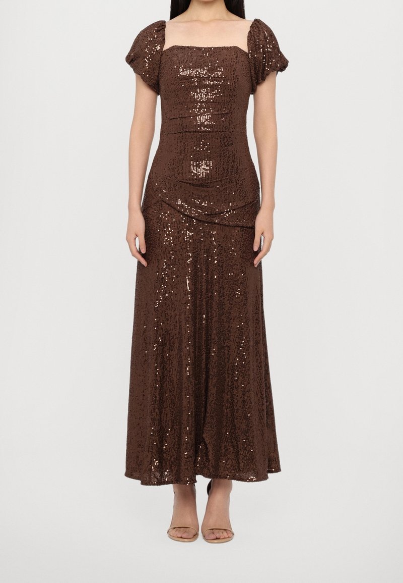 Femme portant une longue robe marron à sequins avec des manches courtes bouffantes et un décolleté carré, debout, les bras relâchés le long du corps.