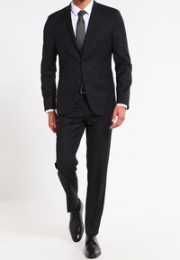 Traje negro con una chaqueta a medida y pantalones a juego. Presenta una camisa blanca y una corbata oscura con lunares. Zapatos de cuero negros.