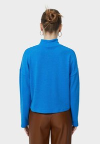 Maglione a coste di un blu elettrico con colletto alto, spalle scese e vestibilità rilassata, abbinato a pantaloni marroni a vita alta.