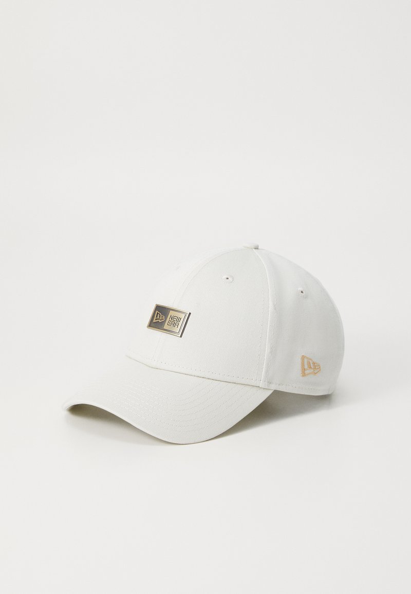 Hvit baseballcap med en strukturert design, utstyrt med et gullmerke og detaljer. Laget av teksturert stoff. Buet skjerm og justerbar stropp.