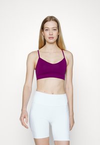 Sutiã desportivo roxo com alças finas, apresentando uma textura suave e um design minimalista. Combinado com calções atléticos brancos de cintura alta.