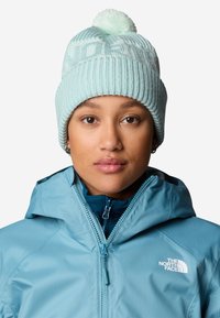 Gorro de punto azul claro con pom-pom, que presenta un ribete vertical y un patrón geométrico. Lleva un abrigo azul a juego.