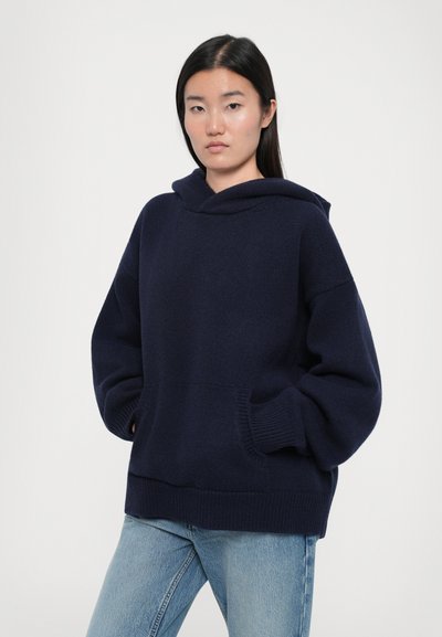 Navy blue oversized hoodie made of knitted fabric, featuring a front pocket, ribbed cuffs, and a relaxed fit. Paired with light blue jeans.