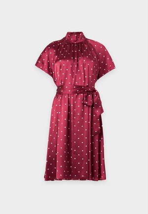 Le mannequin se tient devant un fond uni, vêtue d’une robe bordeaux à pois et de chaussures noires à talons, avec un sac à main noir.