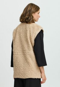 Beige ärmellose Weste mit einer strukturierten, flauschigen Außenseite und einem glatten Stoffbesatz an den Schultern. Vielseitiges Design, das sich gut zum Layering eignet.