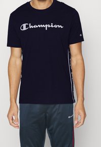 Námořnická modrá bavlněná tričko s krátkými rukávy, které má na přední straně velké bílé logo "Champion" a branding na bocích.