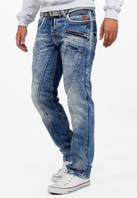 Jeans in denim blu con una finitura usurata, dotati di tasche con zip, cuciture a contrasto e una vestibilità affusolata, abbinati a sneaker bianche.