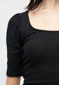 Γυναίκα που φοράει ριμπ πλεκτό μαύρο τοπ με φουσκωτά μανίκια μέχρι τον αγκώνα και τετράγωνη λαιμόκοψη μπροστά σε απλό φόντο.