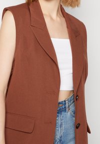 Brun ärmlös blazer med strukturerade slag, enkel knappstängning och sidofickor. Bärs över en vit topp och denimshorts.