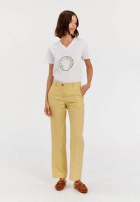 T-shirt blanc à col en V avec un logo vert circulaire, associé à un pantalon droit jaune. Des chaussures slip-on marron viennent compléter la tenue.