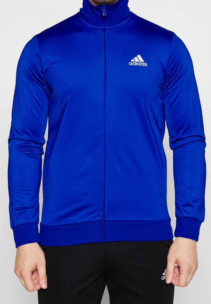 Homme portant une veste zippée bleue Adidas avec un col montant et un pantalon noir Adidas, debout devant un fond uni.