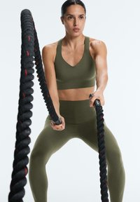 Žena v olivově zeleném sportovním oblečení provádí cvičení s battle rope s výrazem soustředění na obličeji, na jednobarevném pozadí.