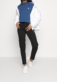 Biała kurtka puchowa, niebieska krótka bluza z kapturem z logo, czarne spodnie z wysokim stanem i guzikami, białe trampki z akcentami.