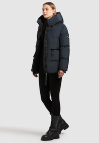 Puffer jacket in dark navy, featuring a high collar, zippered front, two front pockets, and a glossy texture. Paired with black leggings and boots.