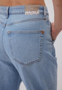 Vaqueros de denim azul claro con un corte relajado, que presentan bolsillos traseros y costuras naranjas. La etiqueta muestra "mavi" y "all blue".