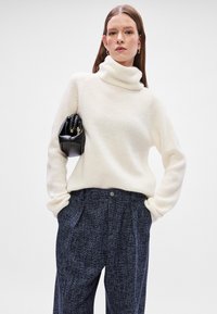 Suéter de gola alta creme com textura canelada, combinado com calças padrão azul-escuro. Modelo segura uma pequena mala preta com alça.