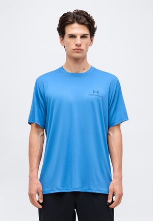 Mladý muž s krátkymi tmavými vlasmi, oblečený v modré tričko Under Armour a čierne nohavice, stojaci tvárou vpred s neutrálnym výrazom.