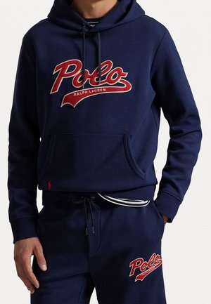 Mężczyzna w granatowej bluzie z kapturem i spodniach dresowych z czerwonym logo "Polo Ralph Lauren", z częściowo widocznymi rękami, na jednolitym tle.