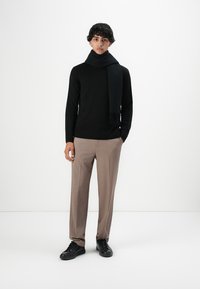 Jonge man in een zwarte trui, zwarte sjaal, beige broek en zwarte schoenen die tegen een effen witte achtergrond staat.
